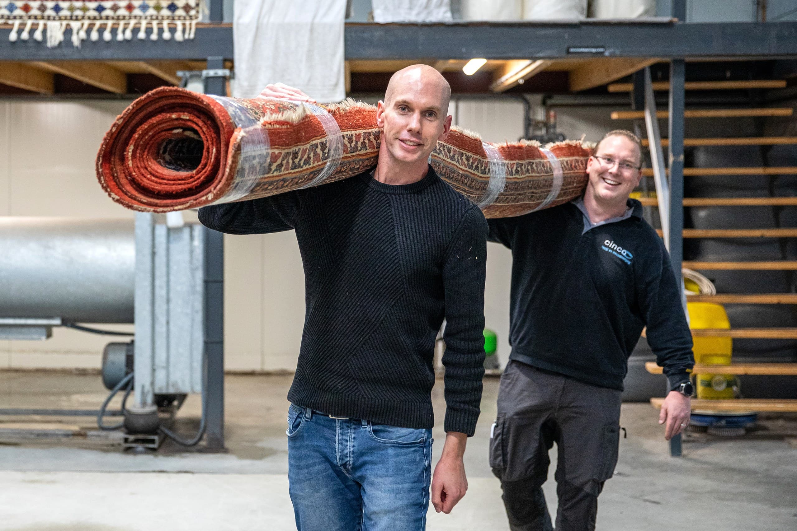 Twee medewerkers dragen een opgerold Perzisch vloerkleed door de reinigingswerkplaats